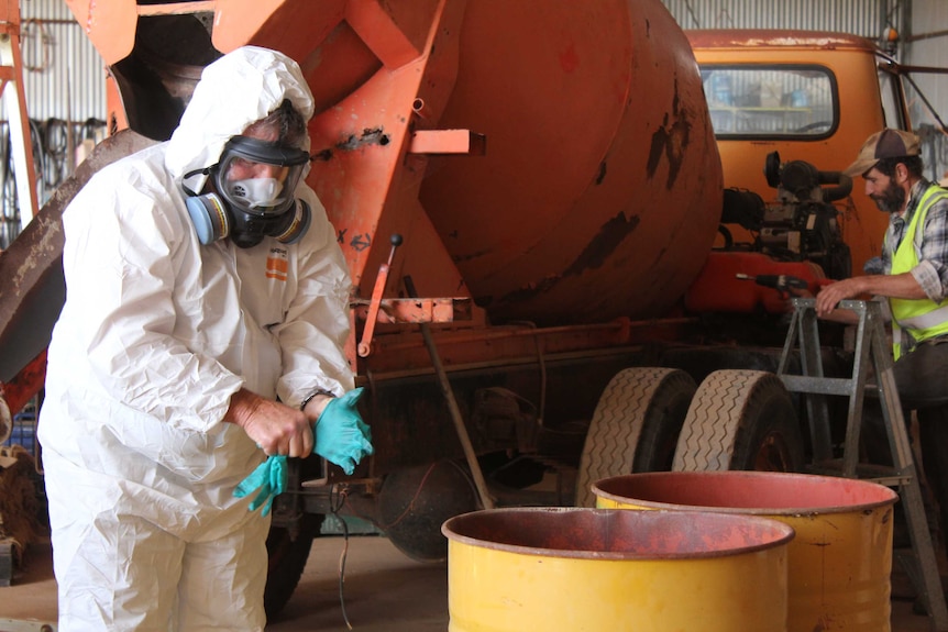 A man in a white chemical hazard suit pulls on gloves in front of an orange cement mixer.