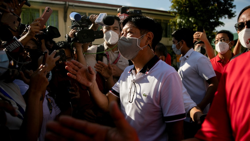 Ferdinand Bongbong Marcos Jr, wearing a face mask, gestures as he walks through a crowd of cameras.