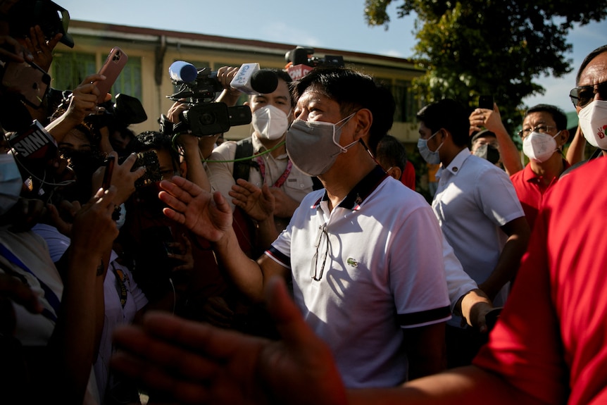 Ferdinand Bongbong Marcos Jr, wearing a face mask, gestures as he walks through a crowd of cameras.