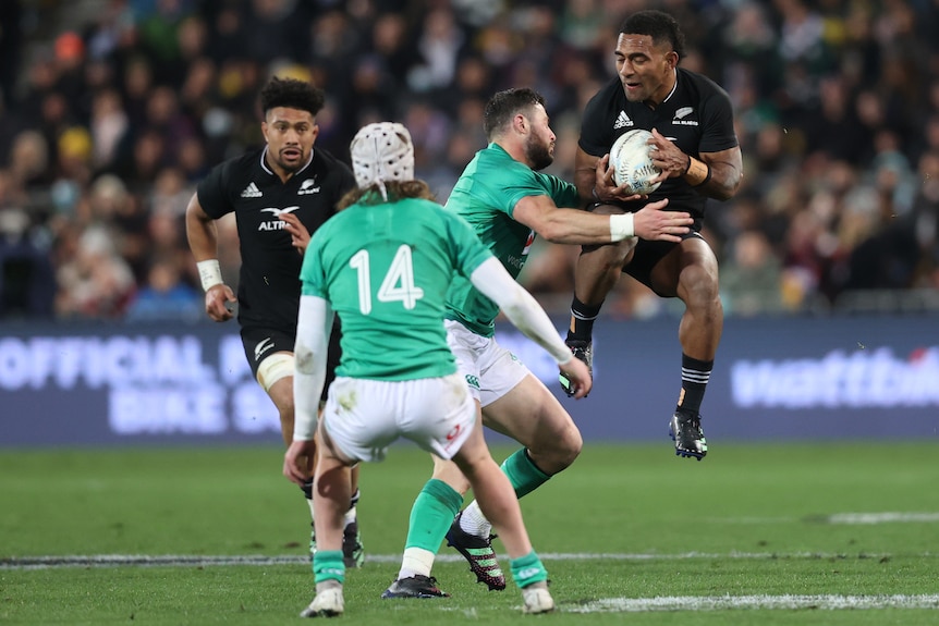 An All Blacks player jumps to catch the ball as an Ireland opponent prepares to tackle him.