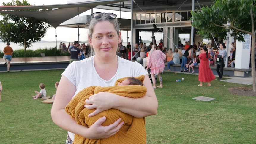 A woman in a white shirt holding a baby in a blanket standing on grass in front of a crowd