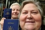 Two women sit on a plane, holding up their passports.