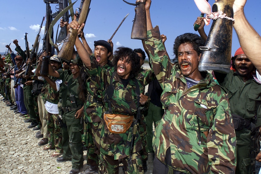 A row of men in camo gear hold up their rifles 