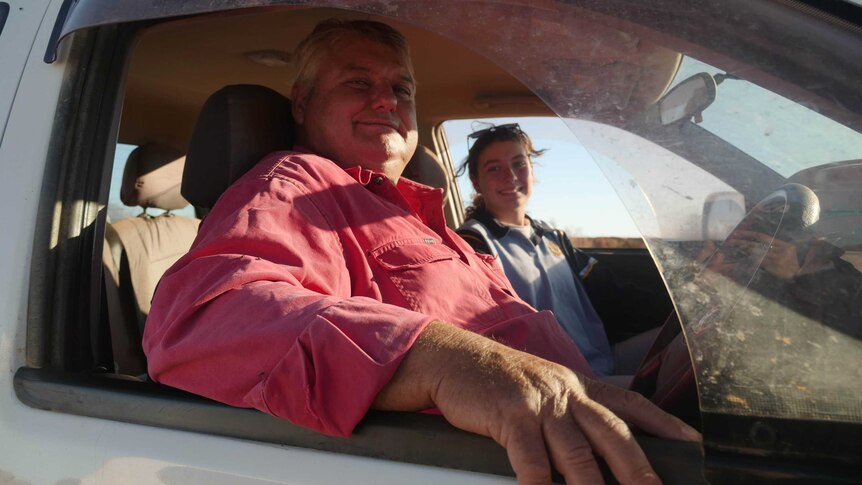 A man and a young girl look at the camera from a car window.