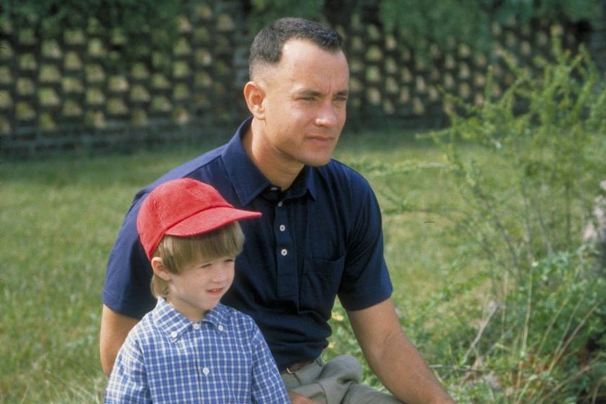 Forrest Gump Jnr holds a lunch bag with sitting on a log with his father
