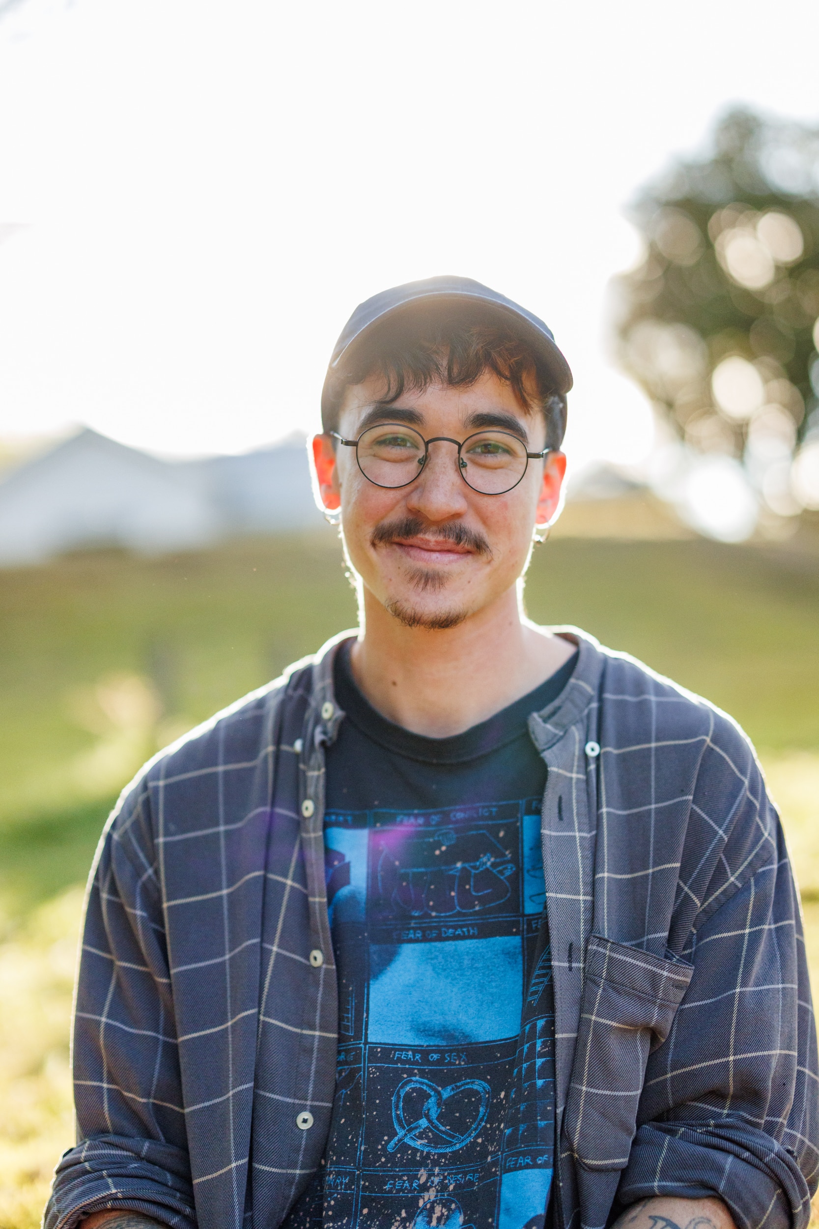 Lee Lai, con bigote y perilla, con gafas, gorra y camisa de cuadros; El sol brilla a través de un árbol de caucho detrás de ella.
