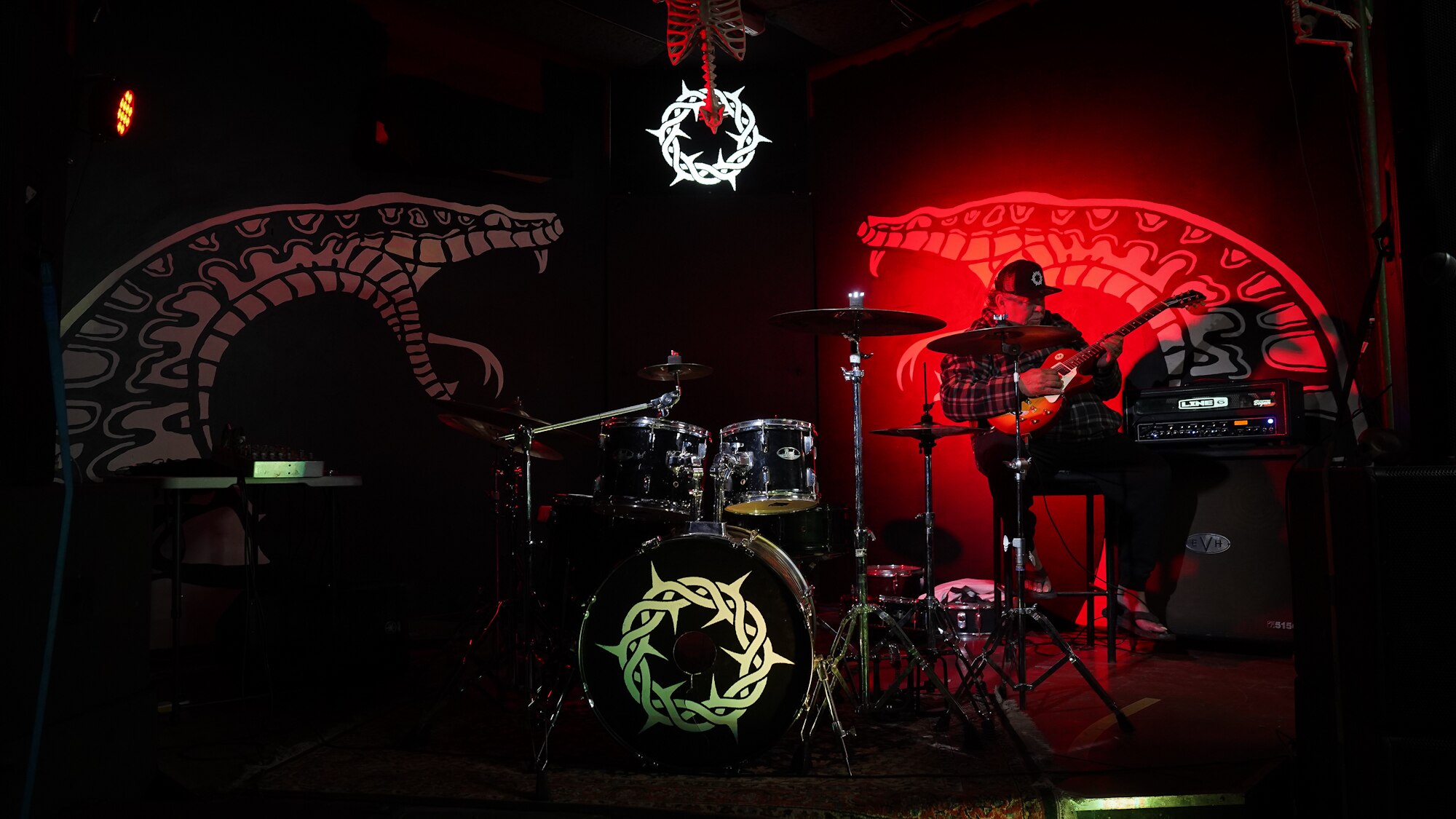 A man sits behind a drum kit playing an electric guitar with a snake lit up by a red light behind him.