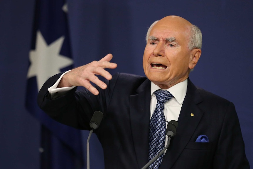 John Howard, eyebrows knitted together, sticks his right hand out while speaking at a microphone in front of an Australian flag.
