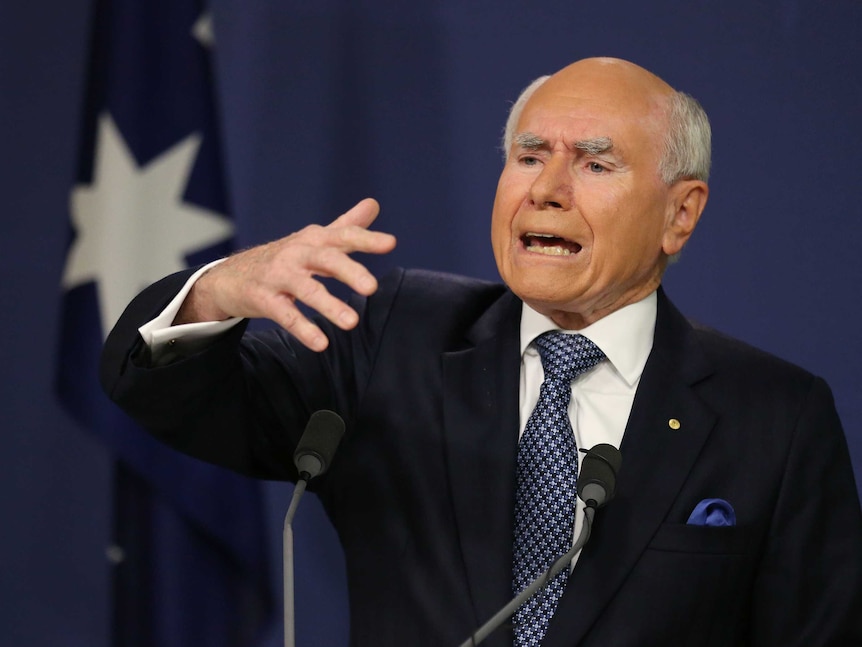 John Howard, eyebrows knitted together, sticks his right hand out while speaking at a microphone in front of an Australian flag.