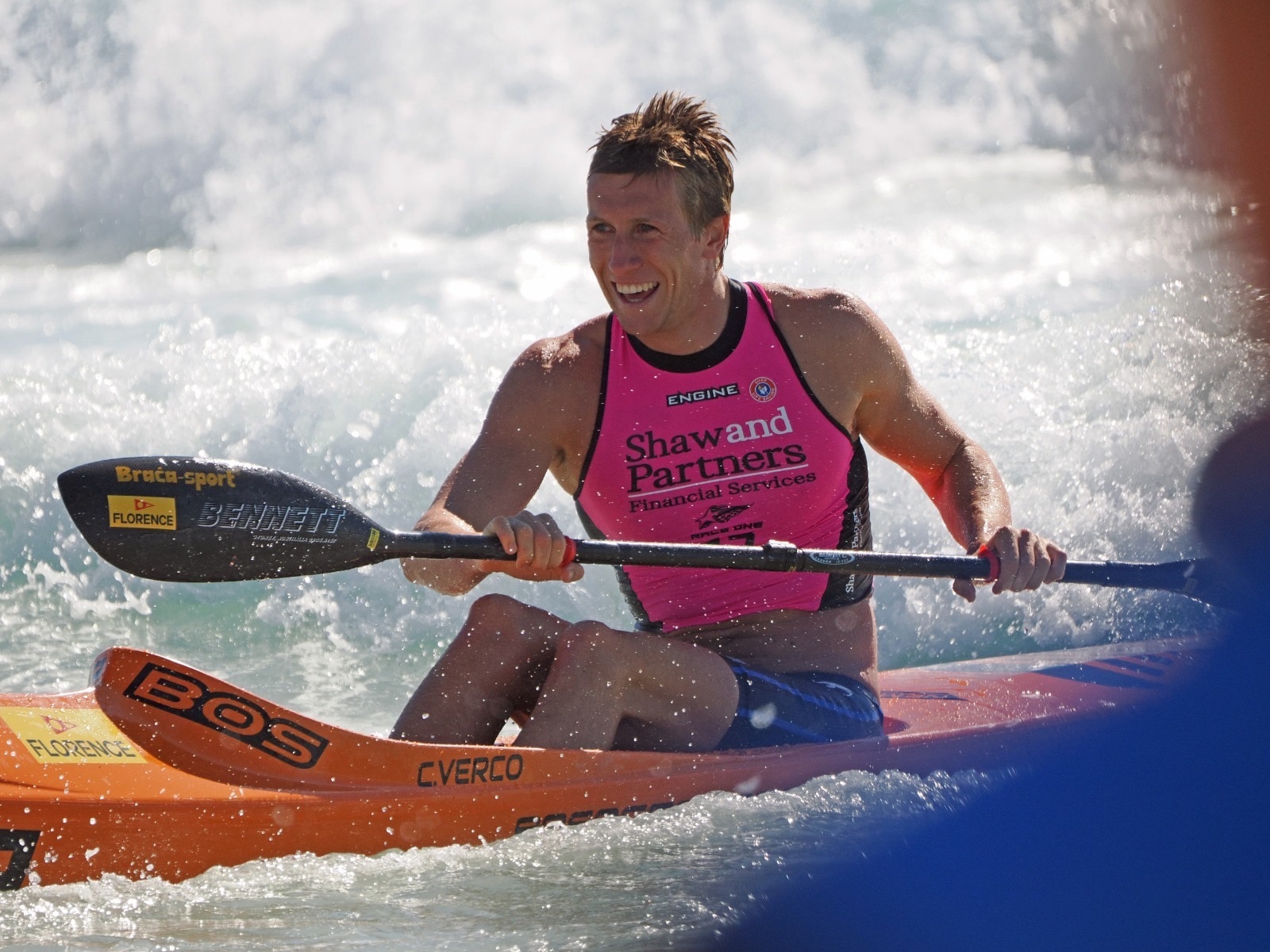 Mr Verco smiling as he uses one big oar to propel himself in a thin flat board forward in waves.