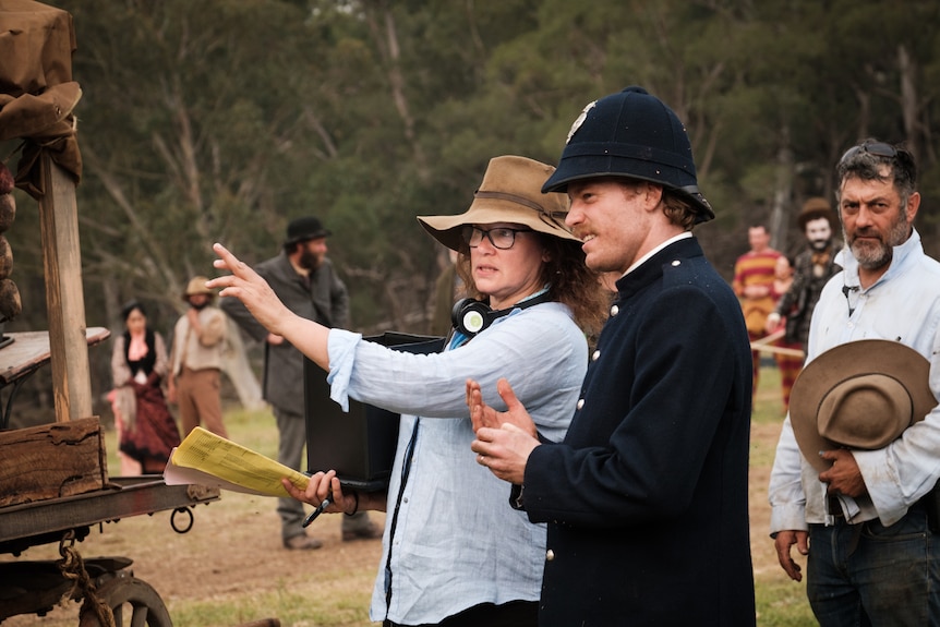 a woman in blue button up and glasses points to distance next to actor wearing an Australian pioneering police uniform