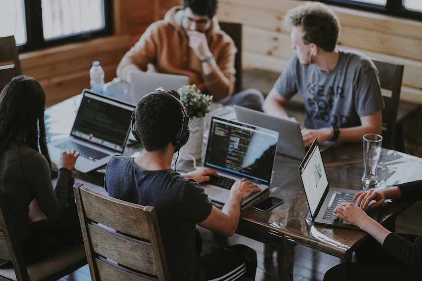 A group of people sit at a table working on laptops and talking.