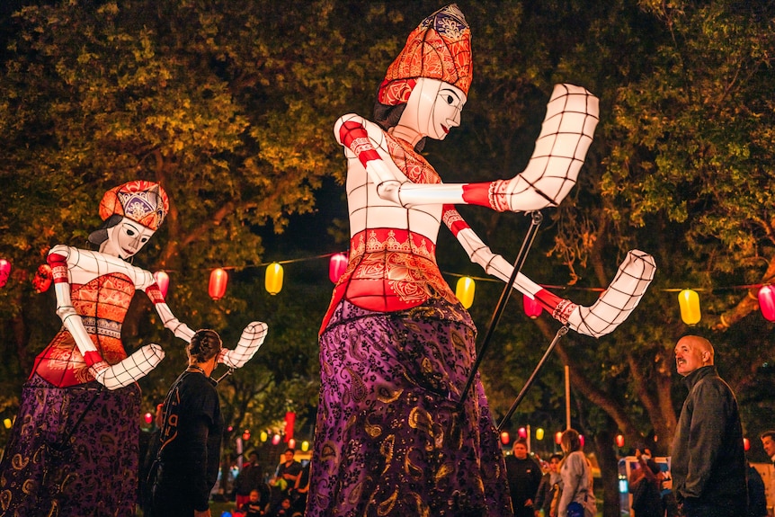 A middle-aged man looks up at two lit-up balloons shaped like giant, regal women in dresses