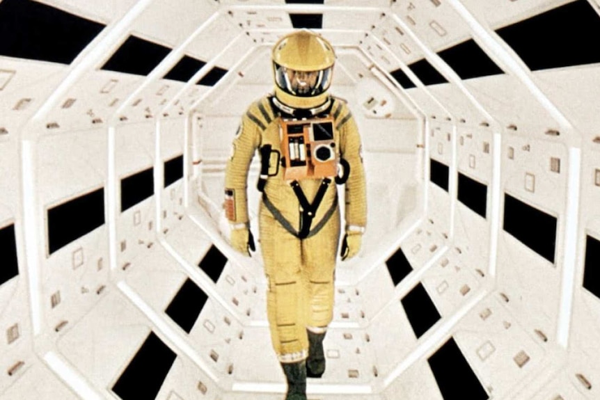 A man in a yellow space suit walks down a white and black corridor 