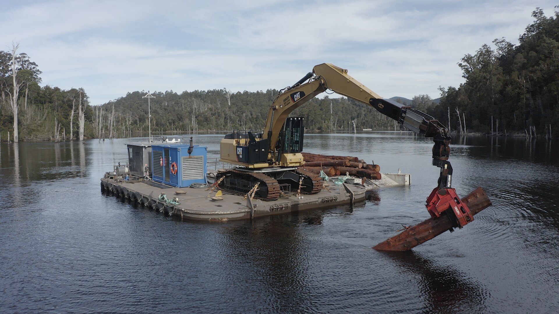 Excavator sits on a floating platform on a large lake.  Excavator is pulling a large piece of timber out of the water. 
