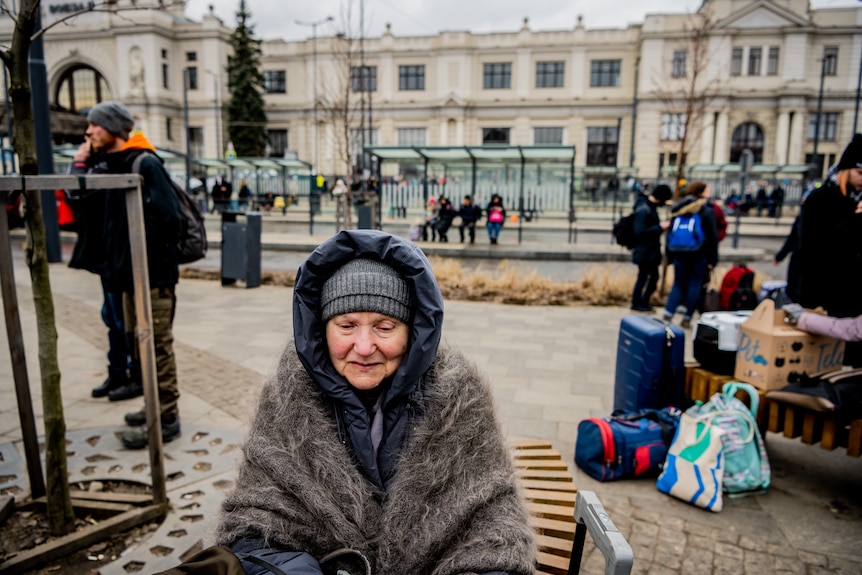 An elderly lady sits out the front of the railway station, her face wrapped in scarves. 