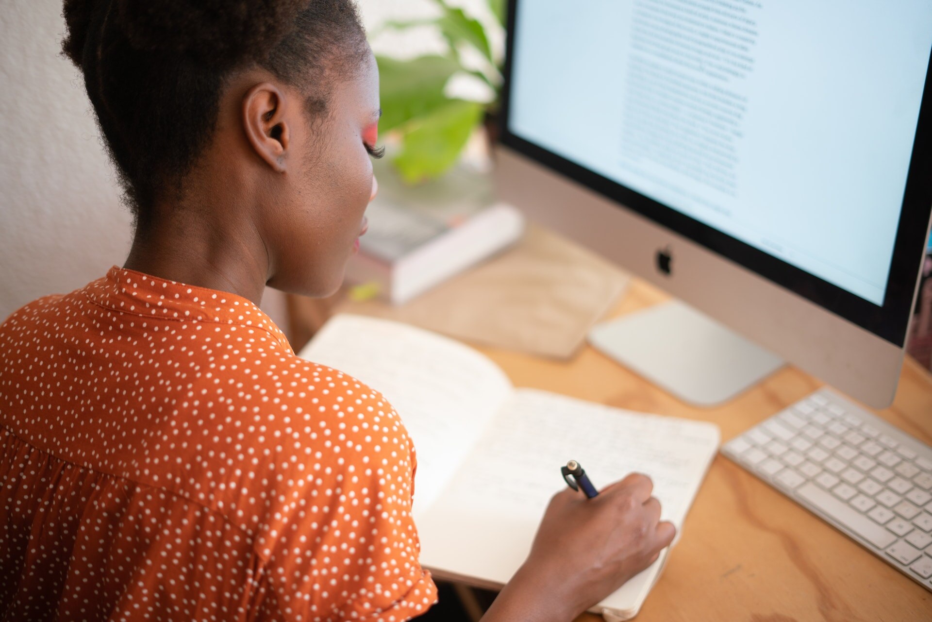 A woman wearing an orange polka dot top writes in a notebook in front of a desktop computer