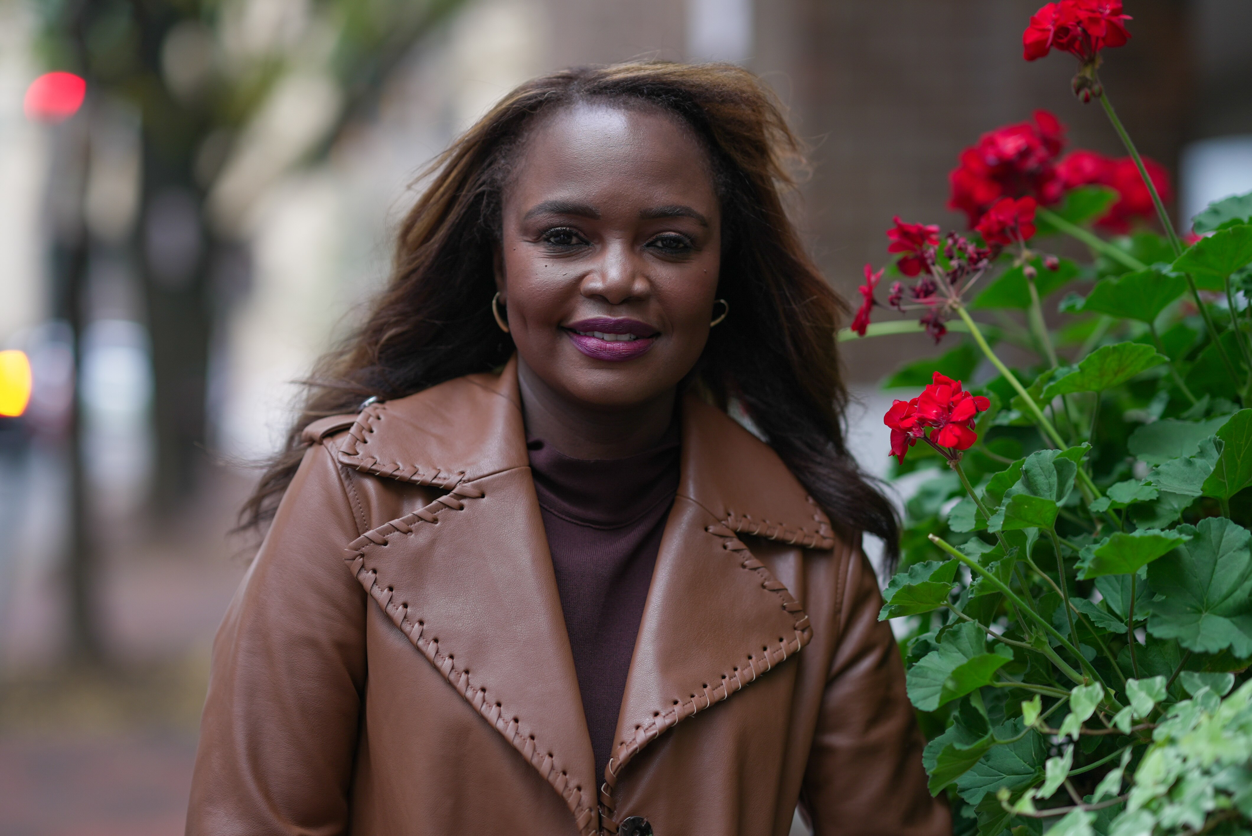 A Haitian woman wearing a brown leather jacket standing next to some flowers.