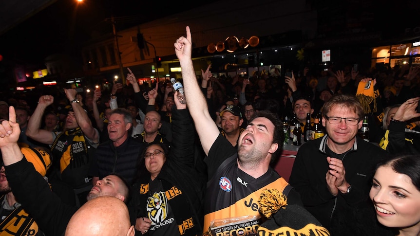 A large crowd of Richmond fans celebrate at night. One man points in the air in celebration.