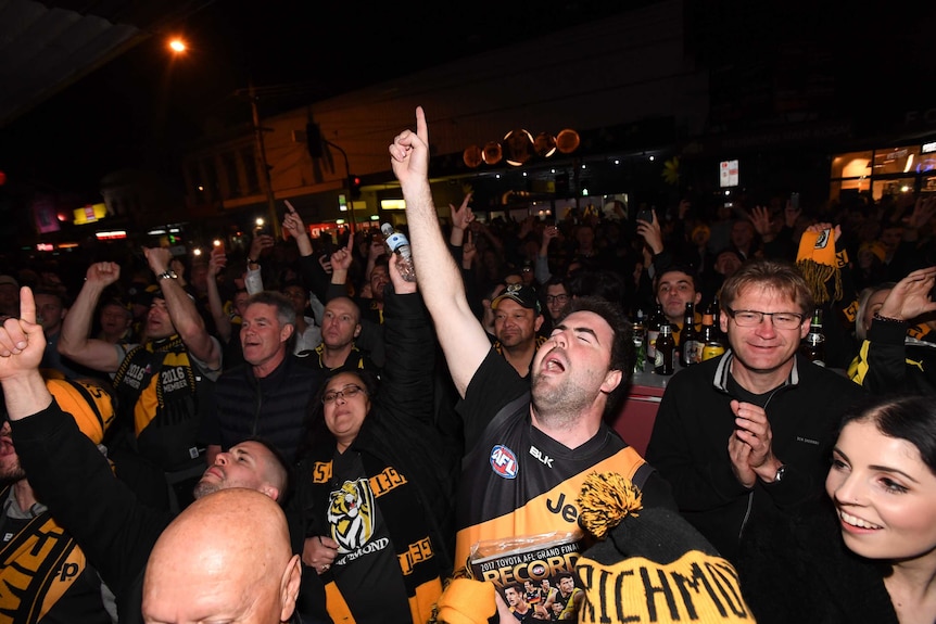 A large crowd of Richmond fans celebrate at night. One man points in the air in celebration.