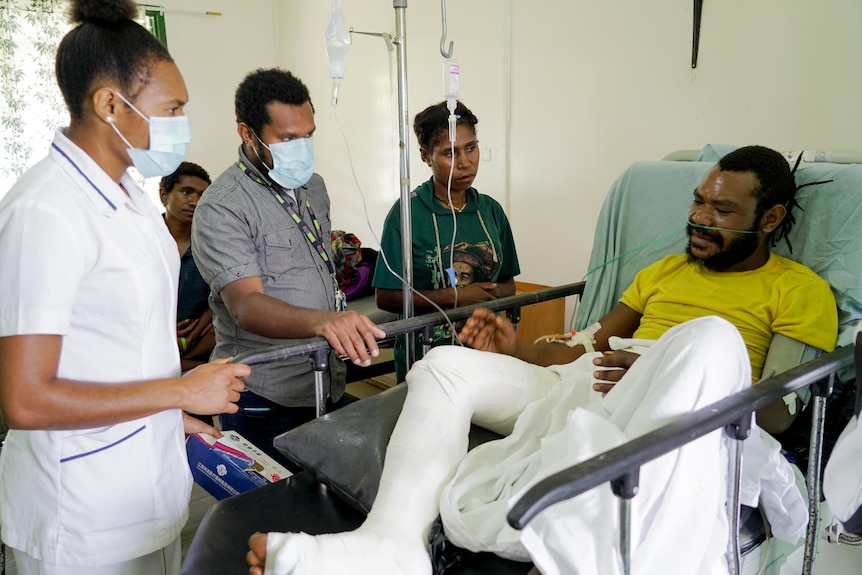 A man with a full leg bandage sits up in a hospital bed, three people stand beside him, two wearing masks