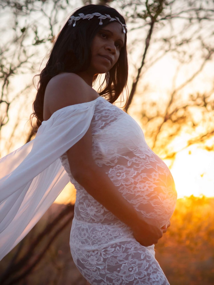 An Indigenous woman wearing a headband poses holding her pregnant belly at sunset in a white lace dress