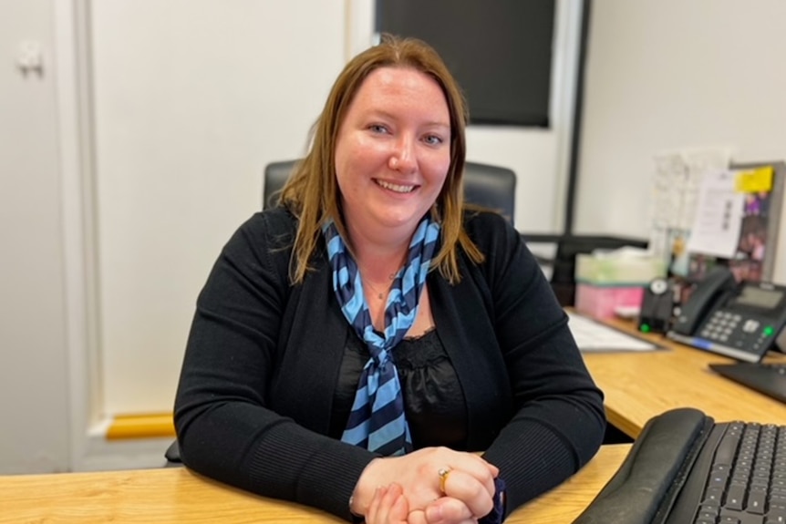 a woman sits at a desk and smiles at the camera. she is wearing black clothing with a blue striped necktie.