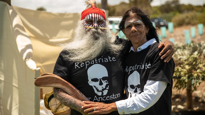 An Aboriginal man with a long grey beard and face paint, holding a boomerang, with an Aboriginal woman with long dark hair