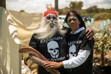 An Aboriginal man with a long grey beard and face paint, holding a boomerang, with an Aboriginal woman with long dark hair