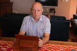 John Allen sitting at a table in front of a television. He is holding a tablet computer in a leather type case.