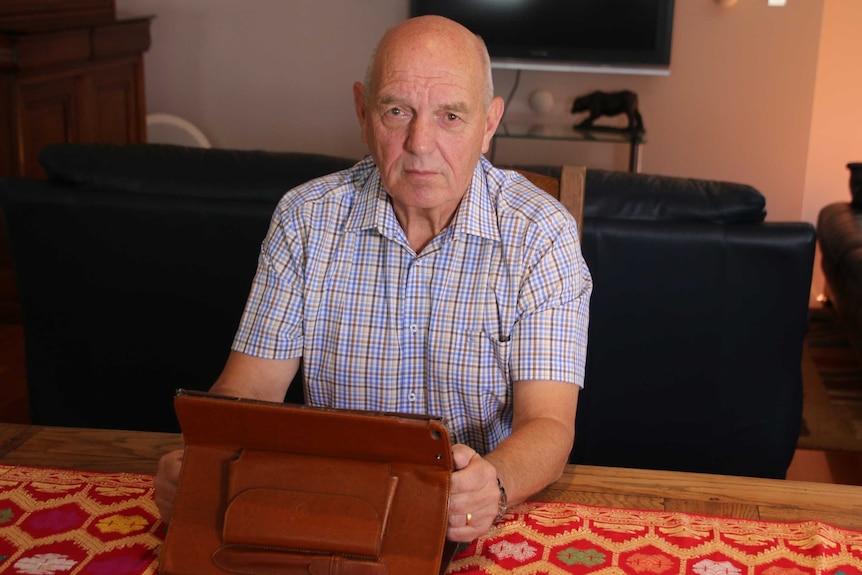 John Allen sitting at a table in front of a television. He is holding a tablet computer in a leather type case.