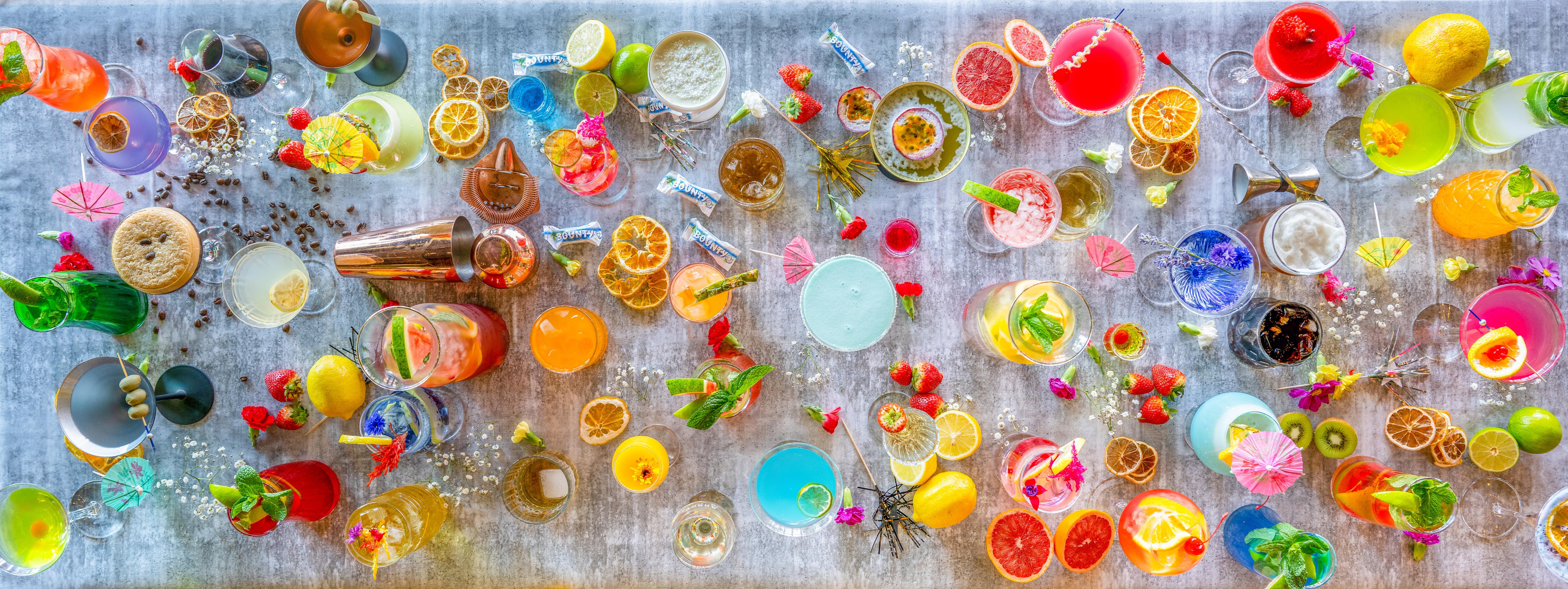 An array of colourful cocktails and flowers is spread across a white table. 