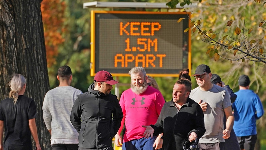Multiple people walk along a busy path in a park. A large illuminated sign is behind them saying 'keep 1.5m apart'