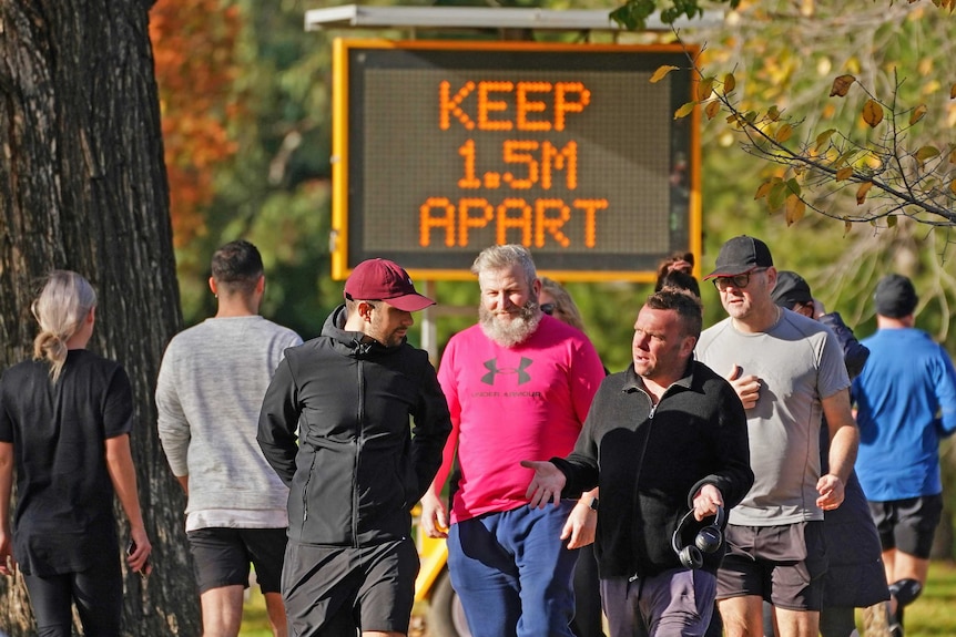 Multiple people walk along a busy path in a park. A large illuminated sign is behind them saying 'keep 1.5m apart'