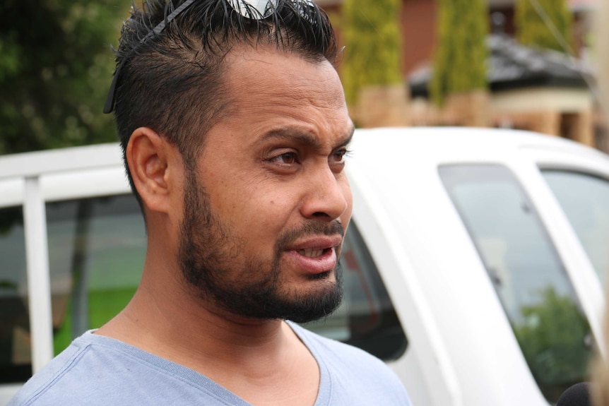 A man with sunglasses sitting up on his head speaks to the media in a suburban street.