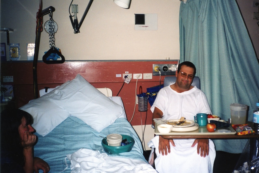 A man wearing a hospital gown sitting next to a hospital bed.