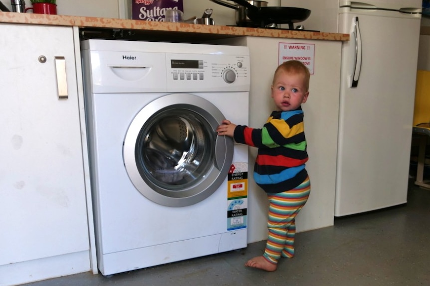 A little boy stands in front of a washing machine inside a shipping container.