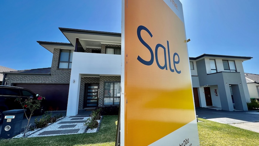 A yellow Sale sign in front of a newly built house on a sunny day.