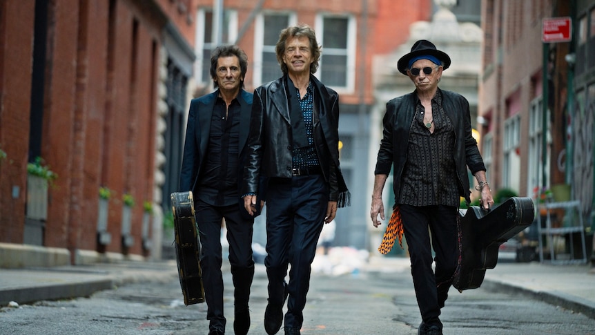 Ronnie Wood, Mick Jagger, Keith Richards of The Rolling Stones walk down a street. Wood and Richards carry guitar cases.
