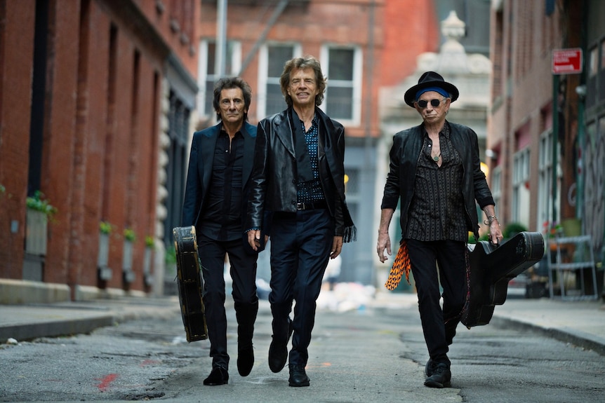 Ronnie Wood, Mick Jagger, Keith Richards of The Rolling Stones walk down a street. Wood and Richards carry guitar cases.