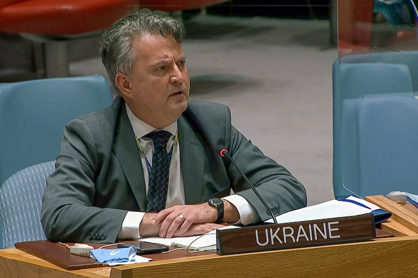A man with grey hair wearing a grey suit is sitting behind a microphone. In front of him is a sign that says Ukraine.