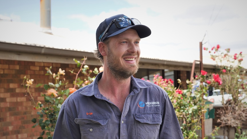 A man in a blue outdoor work shirt wears a cap standing in front of his brick house and rose bushes, smiling.