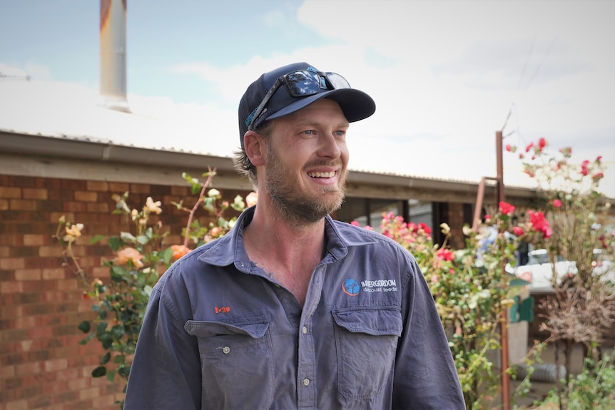 A man in a blue outdoor work shirt wears a cap standing in front of his brick house and rose bushes, smiling.