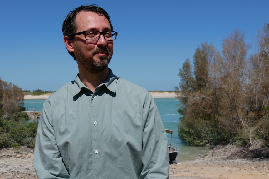 A man with square glasses and a green button up shirt stands in front of a beach
