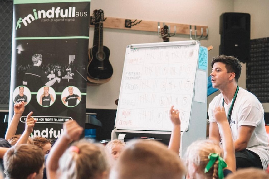 Matthew Runnalls sits to the right next to a whiteboard with a dozen or so children in front of him, some with raised hands.