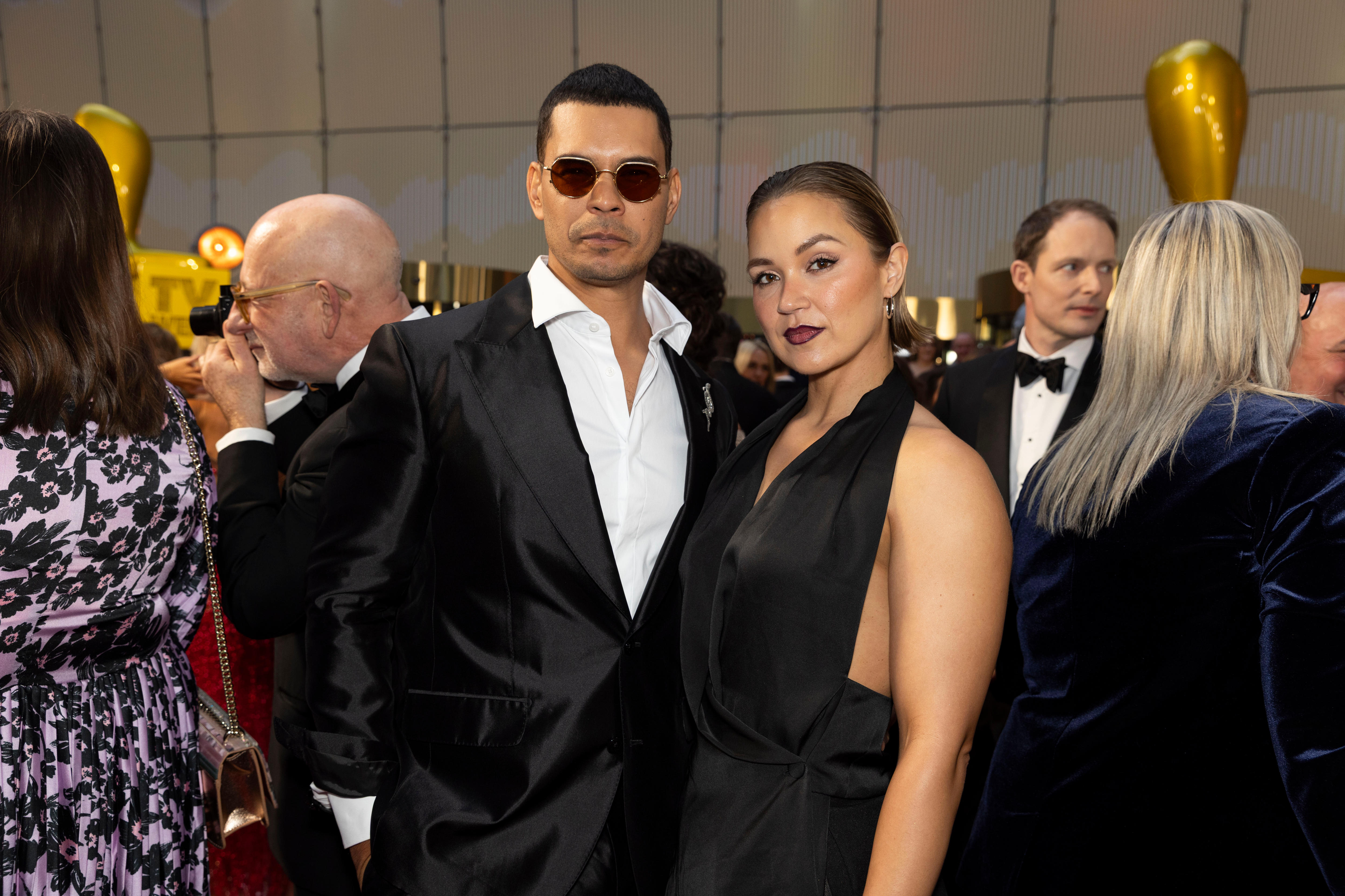 Coles Smith wears a white black suit and aviator sunglasses, Narkle wears a black plunge halterneck dress.