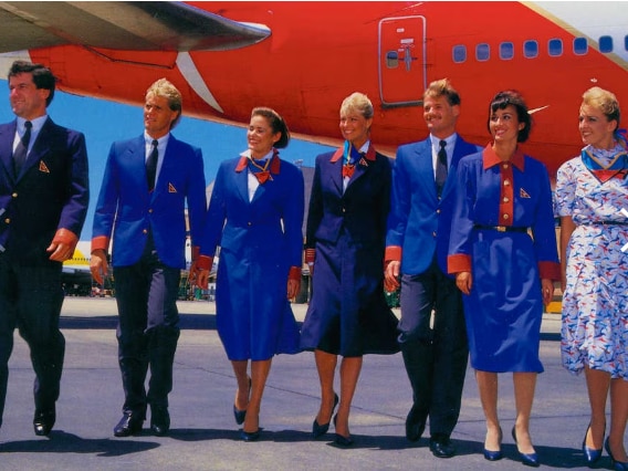An old photo shows seven men and women in various blue and red Qantas uniforms walking in front of a plane.