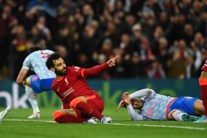 Mo Salah points behind him while half lying on his side with two other players lying next to him