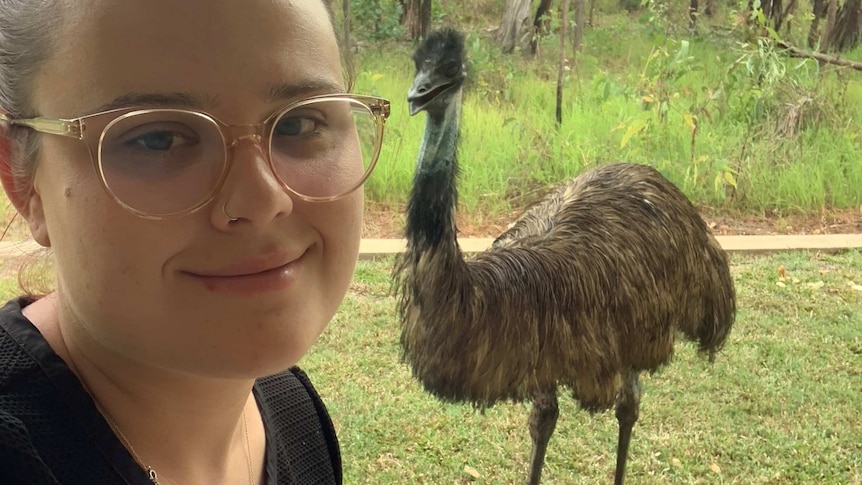 A woman with red hair and glasses smiles at the camera. She is standing outdoors with an emu in the background.