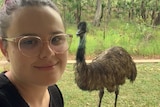 A woman with red hair and glasses smiles at the camera. She is standing outdoors with an emu in the background.